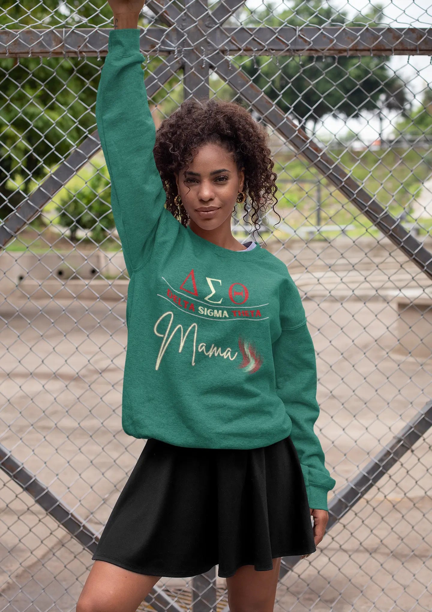 A person stands confidently in front of a chain-link fence, wearing a green Delta Sigma Theta sorority sweatshirt from the Gifts For Mom store and a black skirt. The background features blurred greenery and concrete.