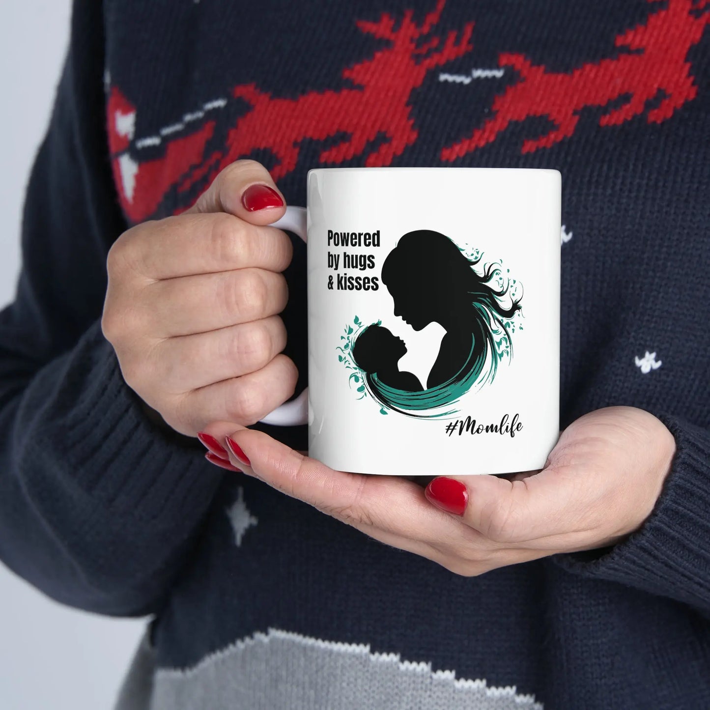 A person with red nail polish holds a coffee cup from the Gifts For Mom store, featuring a silhouette of a woman and child alongside the text Powered by hugs & kisses. The hashtag #Momlife is below the image. The person is wearing a sweater with reindeer designs.