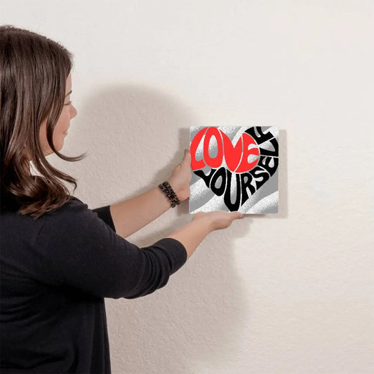 A person is hanging a stylized heart shape formed by the words Love Yourself in bold letters. Love is in red, and Yourself is in black, set against a background of wavy gray and white stripes. Ideal as metal wall art from our Gifts for Mom store, its a heartfelt reminder to cherish oneself.