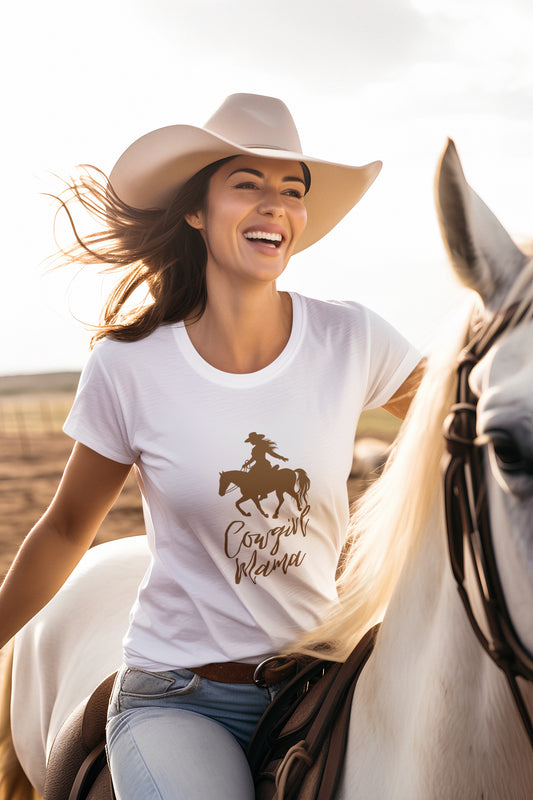 A woman in a cowboy hat and a white Cowgirl Mama T-shirt, from the Gifts For Mom store, is joyfully riding a white horse in the sunny, open field.