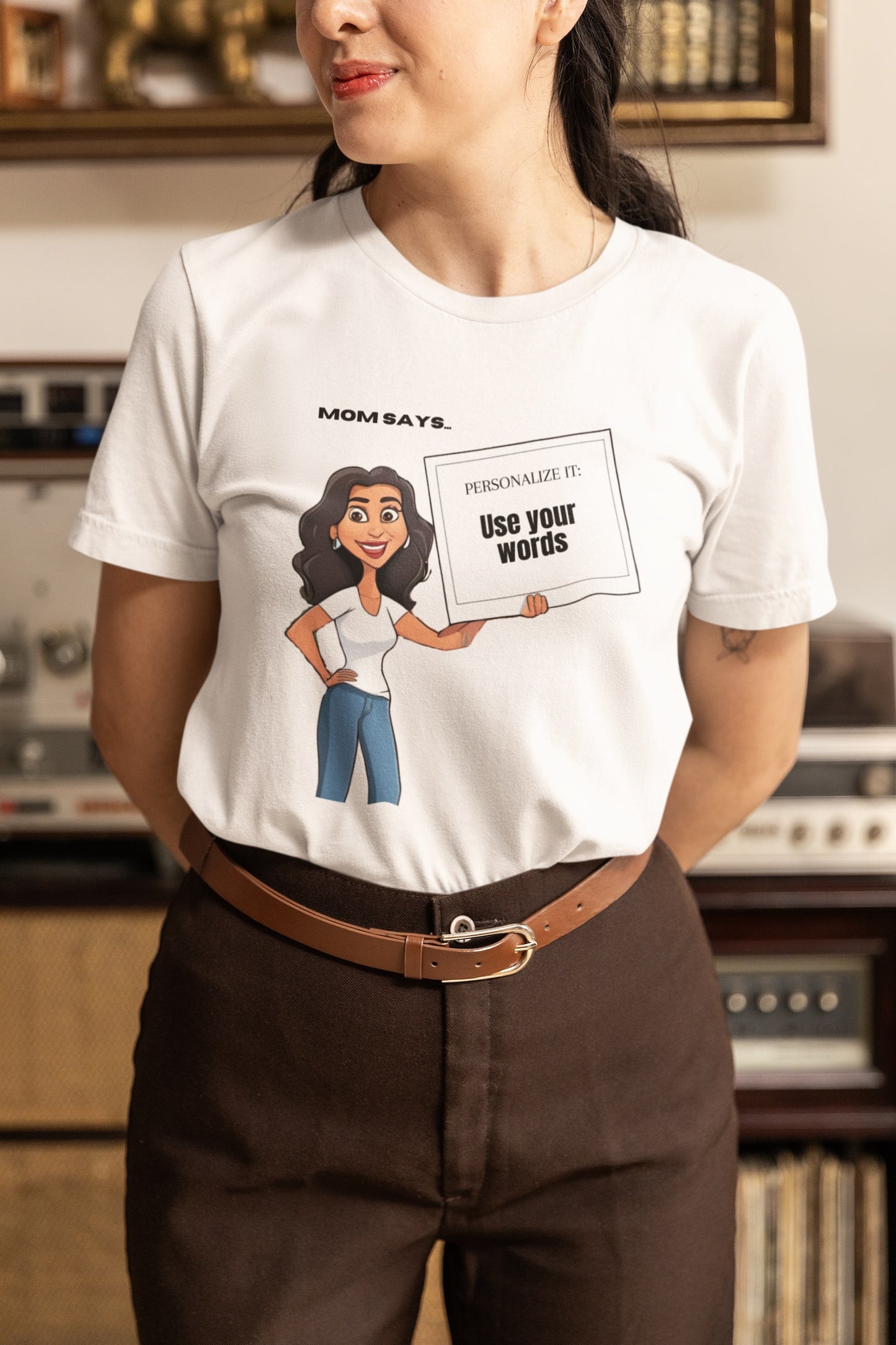 A woman sports a personalizable white t-shirt featuring a cartoon of a woman holding a sign that declares, Mom says: Use your words. Shes paired it with brown pants and a belt. The scene is set against vintage audio equipment, highlighting the charm of personalizable tees from the Gifts For Mom store.